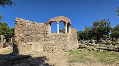 Ruined stone rotunda with brick arches stands among scattered ancient stones and trees.