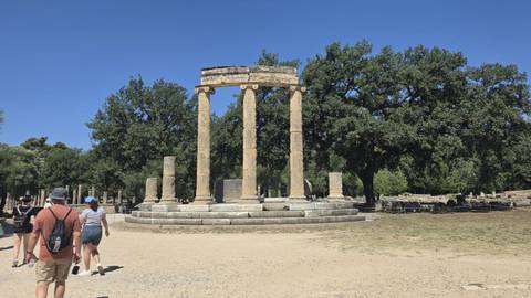 Circular arrangement of ancient columns known as the Philippeion stands in a sandy clearing with tourists nearby.
