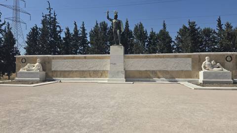 Statue of King Leonidas stands at the Thermopylae monument flanked by marble relief walls and cypress trees.