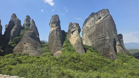 Series of tall, narrow Meteora rock pinnacles rise against a clear blue sky above green forest.