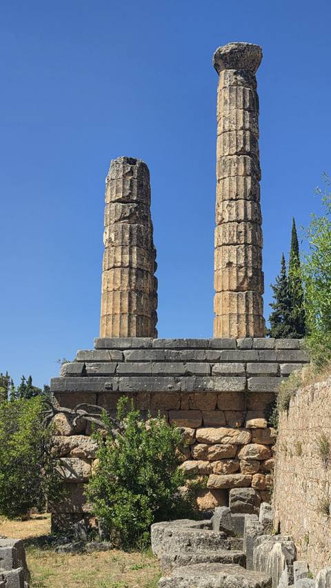Two weathered stone columns rise on a stepped base against a clear blue sky at Delphi.