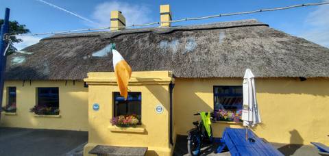 Sunny yellow thatched-roof cottage adorned with Irish flag, flower boxes and blue picnic bench