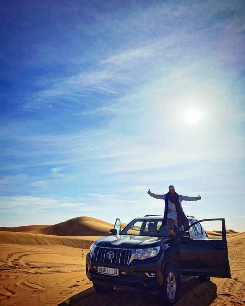 Traveller standing on a 4x4 roof giving thumbs up beneath vast blue desert sky and bright sun