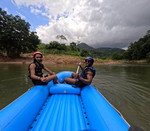 Two rafters in helmets relax on an inflatable blue raft floating down a jungle river under moody clouds.