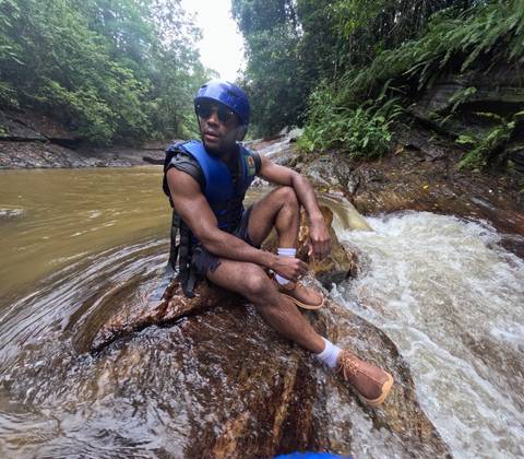 Adventurer in helmet and life vest sitting on a wet rock beside a rushing jungle stream.