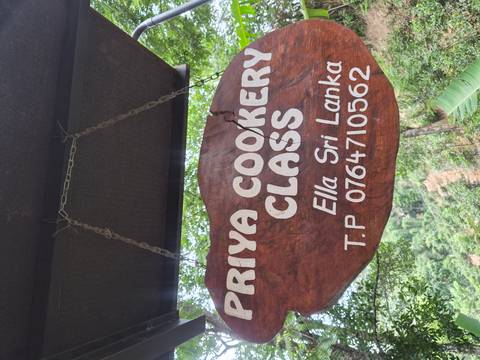 Wooden hanging sign reading 'Priya Cookery Class, Ella Sri Lanka' surrounded by lush greenery.
