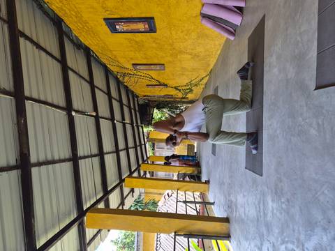 Practitioner holding a yoga pose on a mat in a shaded open corridor with yellow pillars.
