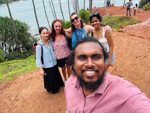 Group selfie of smiling travelers on a red dirt cliff overlooking Sri Lanka’s lush coastline and palms.
