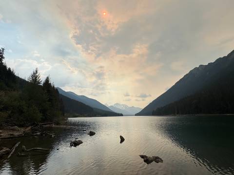 Tranquil alpine lake flanked by forested slopes and distant snowy peaks under pastel skies.