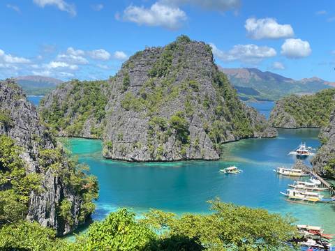 Spectacular limestone cliffs encircling an emerald lagoon with outrigger boats floating on vivid turquoise water under bright blue skies.