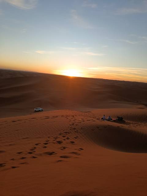 Small group watching a fiery desert sunset from high sand dunes with a lone 4x4 parked below.