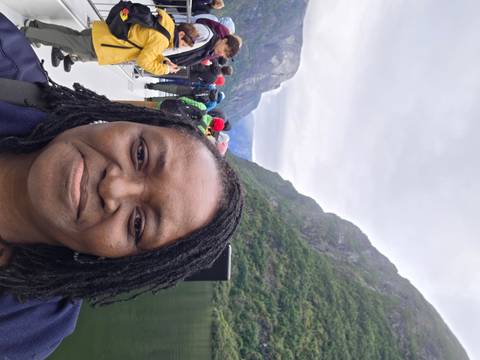 Selfie aboard a boat cruising through a steep green fjord under cloudy skies with other passengers behind.