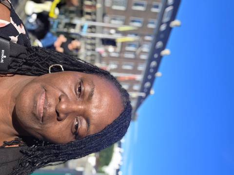 Close-up selfie of a woman on a sunny day with blurred urban buildings in the background.