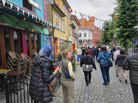 Visitors walk along a narrow cobblestone street lined with brightly painted wooden houses, cafés and flowers.