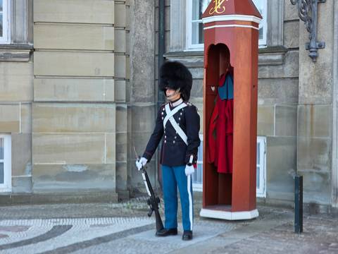 Uniformed royal guard stands at attention beside a red sentry box outside a stone palace wall.