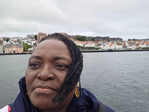 Close-up selfie of a woman on a boat with white seaside houses and grey sky in background.