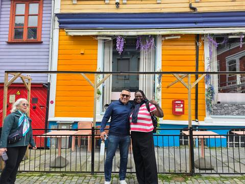 Travelers smile and pose outside a brightly painted wooden façade decorated with flowers.