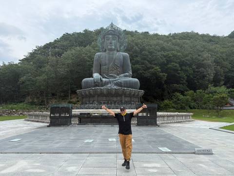 Man posing thumbs up before large seated Buddha statue in forested temple grounds