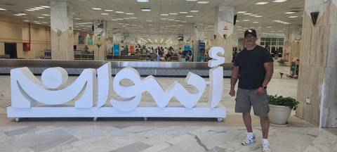 Traveler standing beside large illuminated Aswan sign inside the airport terminal