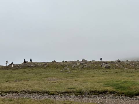 Tiny silhouettes of hikers spread across a foggy, grassy ridge under low clouds.