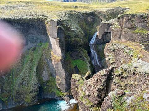 Deep canyon with slender waterfall, partially obscured by a thumb covering the lens edge.