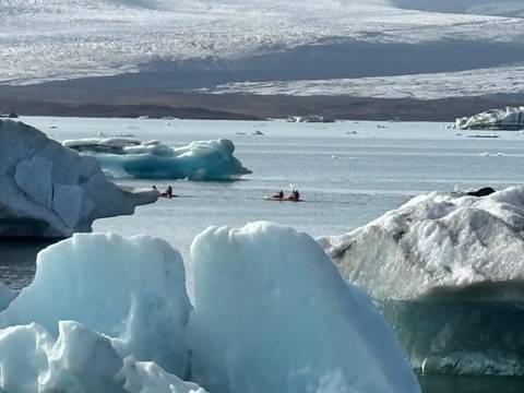 Kayakers paddling among blue icebergs on a glacial lagoon in bright sunlight.