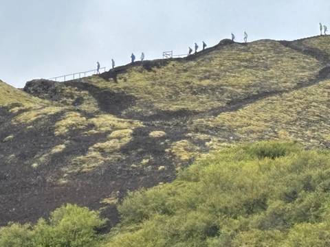 Line of hikers ascending the ridge of a mossy volcanic slope with switchback paths.