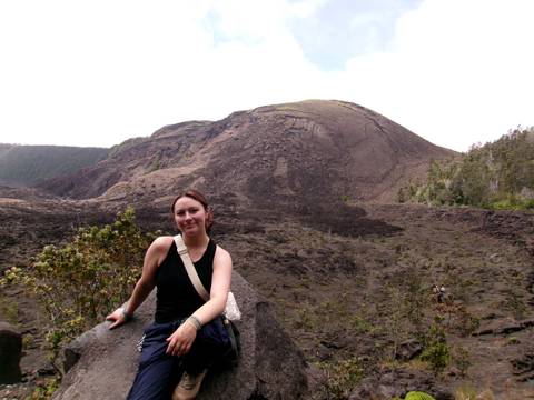 Traveler posing on a volcanic landscape with a broad lava field and cratered hill behind.