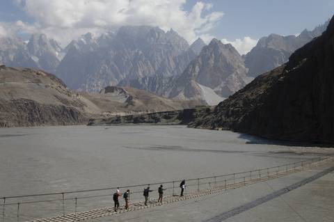 Small group crosses a narrow suspension bridge over a glacial lake amid jagged Karakoram peaks.