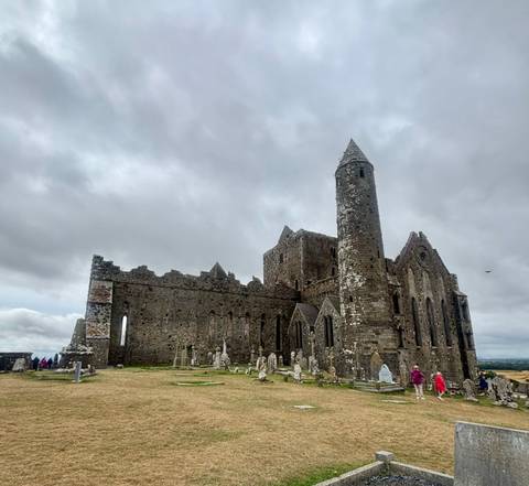 Medieval stone cathedral ruins on a hilltop with graveyard below under grey skies.