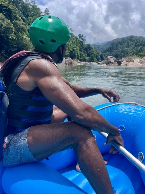 Close-up of a man in a life jacket gripping a blue inflatable raft on a calm river with rocky shore in the background.