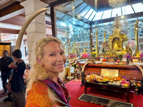 Woman smiles inside an ornate Buddhist shrine hall filled with golden statues, flowers and offerings.