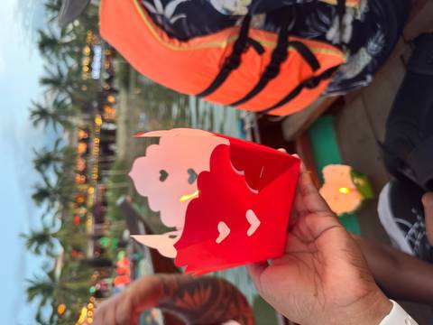 Hand holding a red paper lantern with heart cutouts on a wooden boat during Hoi An river lantern festival.