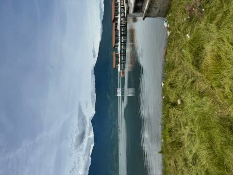 Calm lake with distant pier buildings and ‘Kuri’ sign backed by green mountains under light cloud.