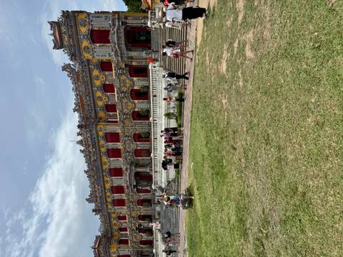 Tourists exploring expansive lawn and ornate façade of Hue Imperial Palace on a cloudy day.