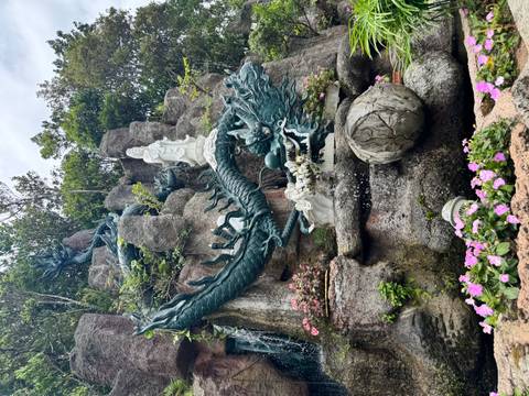 Intricate dragon and deity sculptures in rocky garden fountain with blooming flowers.