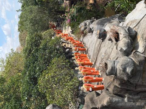 Long procession of orange-robed monk statues lines a rocky garden path beneath leafy trees.