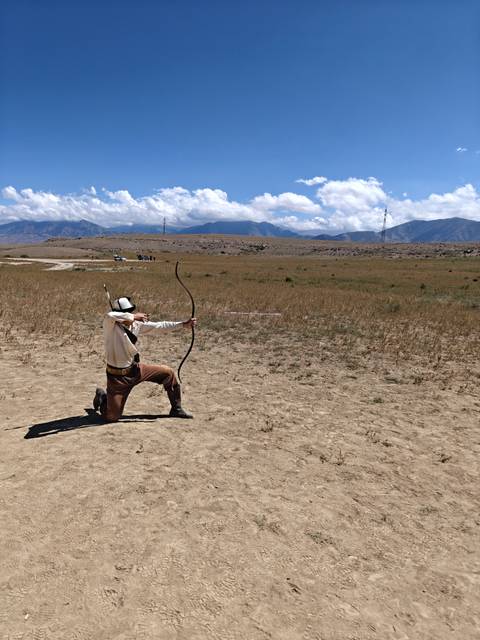 Traditional archer kneels on the steppe drawing a bow toward distant targets against mountain backdrops.