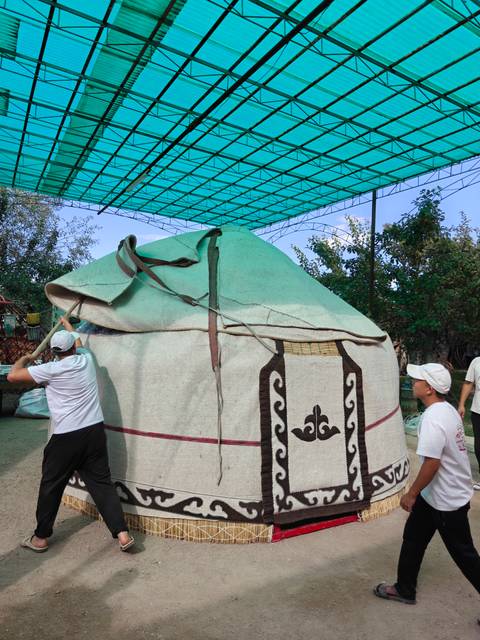 Two men assemble a traditional yurt frame under a shaded structure in a village yard.