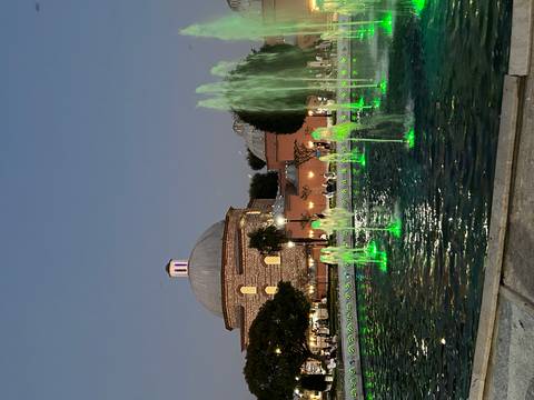 Evening scene of illuminated fountains glowing green before a domed historic bathhouse.