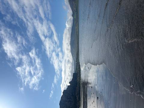 Rippling shoreline meets towering sea cliffs under bright sunshine and scattered clouds.