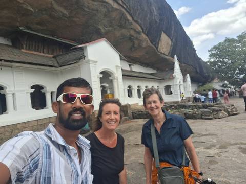 Guide and two visitors take a selfie in front of white-arched buildings set beneath an overhanging rock temple.