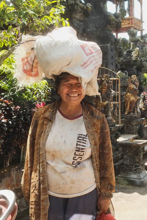Cheerful Balinese woman smiles broadly while balancing a burlap sack on her head.