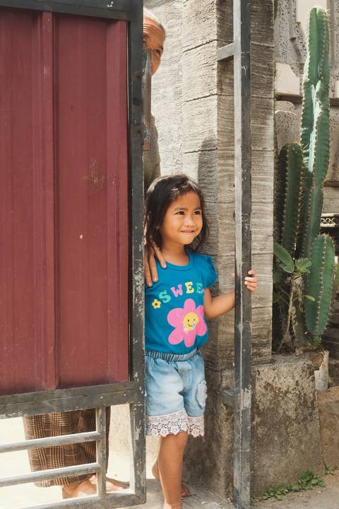 Smiling young girl peers from a doorway beside tall cactus plants in a Balinese village.