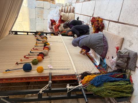 Women sitting on cushions hand-weaving a large carpet using colorful wool in a traditional workshop.