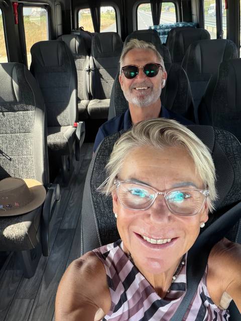 Close selfie of two travelers seated inside a tour van with rows of empty seats behind them.