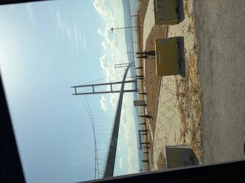 Long-span suspension bridge crossing a strait, viewed from a roadside with concrete barriers and dry grass under a bright sky.