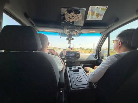 Interior view of a van with two men in the front seats driving along a rural road under sunny skies.