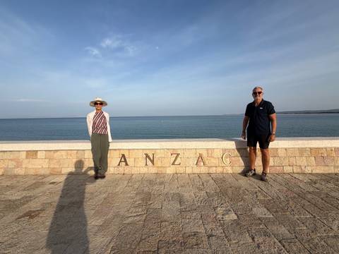 Two travellers pose beside a seaside memorial wall engraved with the word 'ANZAC', calm blue water behind.