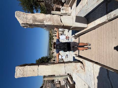 Visitor stretches arms between two tall marble pillars amid ancient stone ruins under a deep blue sky.